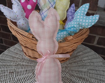 May include: A collection of handmade fabric bunny decorations in a wicker basket. The bunnies are made of various patterned fabrics, including polka dots and plaid, in pastel colors. One pink plaid bunny is in front, tied with a pink ribbon. The basket is on a white wicker surface.