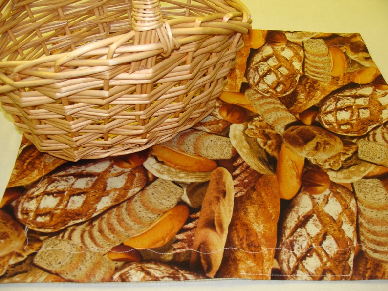 May include: A woven wicker basket sits on a fabric placemat. The placemat features a repeating pattern of various types of bread, including baguettes and loaves, in shades of brown and tan. The basket is light brown.