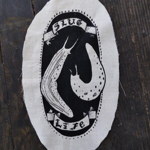 May include: An oval-shaped off-white fabric patch featuring a black screen-printed design of two slugs inside a dotted border. A banner at the top reads SLUG and a banner at the bottom reads LIFE in a stylized black font.