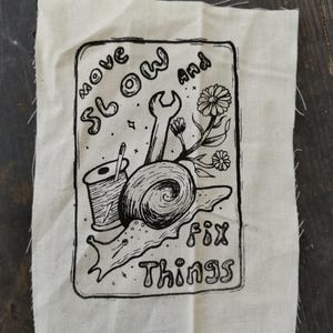 May include: A rectangular piece of off-white fabric featuring a black ink illustration of a snail, a spool of thread with a needle, a wrench, and flowers. The text "move slow and fix things" is printed in a stylized font within the rectangular border.