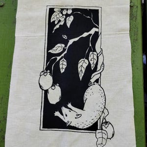 May include: A rectangular off-white fabric tea towel featuring a black ink block print. The design depicts an opossum hanging from a branch with leaves and round fruits, all enclosed within a thin black rectangular border frame.