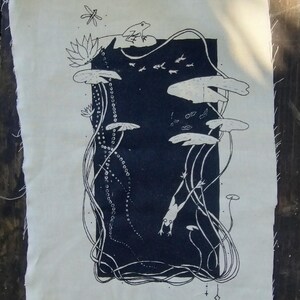 May include: A rectangular piece of off-white fabric featuring a dark navy blue central panel with a black ink illustration of a pond scene. The design includes frogs, lily pads, lotus flowers, a dragonfly, and small fish, with raw, frayed edges on the fabric.