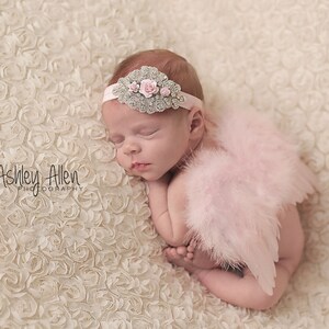 May include: A sleeping baby wearing a pink feathered angel wings costume and a silver headband with pink flowers. The baby is lying on a white, textured background.