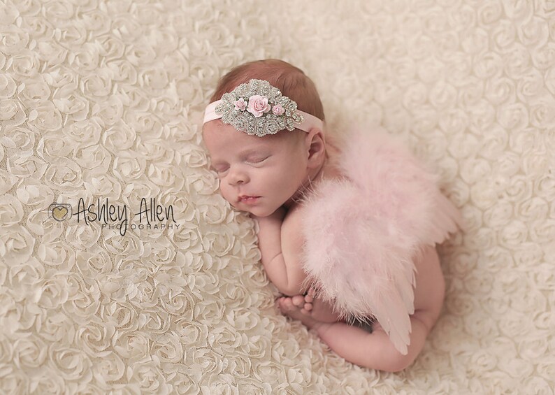 May include: A sleeping baby wearing a pink feathered angel wings costume and a silver headband with pink flowers. The baby is lying on a white, textured background.