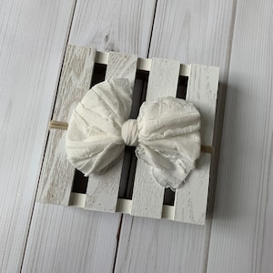 May include: A white, ruffled fabric bow with a knotted centre, displayed on a white wooden crate.
