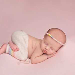 May include: A sleeping baby wearing a white knitted outfit with a rainbow stripe on the leg and a colorful beaded headband.