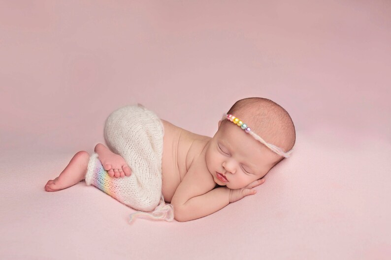 May include: A sleeping baby wearing a white knitted outfit with a rainbow stripe on the leg and a colorful beaded headband.