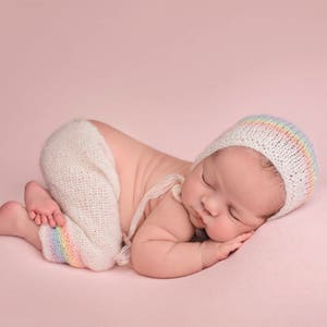 May include: A newborn baby sleeping on a pink background, wearing a white crocheted hat with a rainbow stripe and a white crocheted outfit.