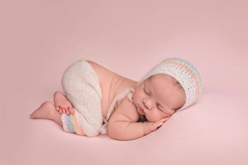 May include: A newborn baby sleeping on a pink background, wearing a white crocheted hat with a rainbow stripe and a white crocheted outfit.