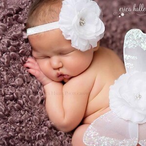 Puede incluir: Un bebé dormido con una diadema floral blanca y un conjunto de alas de mariposa a juego con detalles de purpurina. El bebé está acostado sobre una manta texturizada de color malva. Es un retrato profesional de recién nacido.
