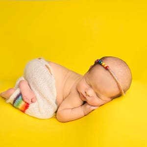 May include: A newborn baby wearing a white knitted outfit with a rainbow stripe detail, sleeping on a yellow background. The baby is wearing a colorful beaded headband.