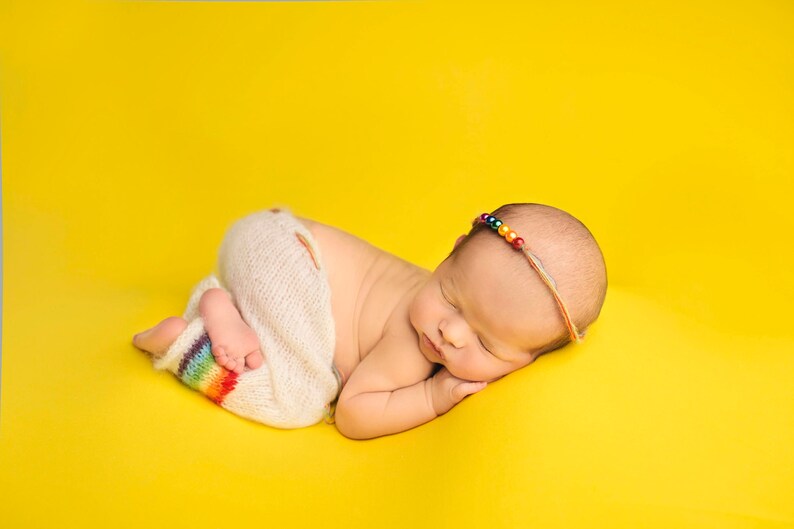 May include: A newborn baby wearing a white knitted outfit with a rainbow stripe detail, sleeping on a yellow background. The baby is wearing a colorful beaded headband.