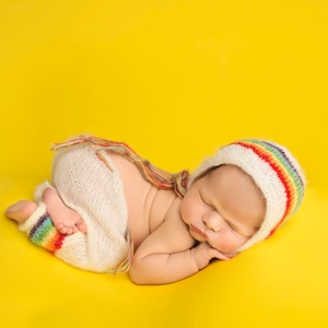 May include: A newborn baby wearing a white crocheted outfit with rainbow stripes and a matching rainbow crocheted hat sleeps on a yellow background.