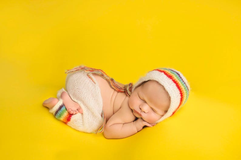 May include: A newborn baby wearing a white crocheted outfit with rainbow stripes and a matching rainbow crocheted hat sleeps on a yellow background.