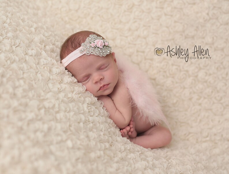 May include: A sleeping baby girl wearing a pink feather boa and a sparkly headband with pink flowers. The baby is lying on a white, fluffy blanket.