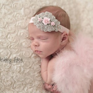 May include: A sleeping baby girl wearing a pink feathered angel wings costume and a silver headband with pink flowers and rhinestones. The baby is lying on a white, textured background.