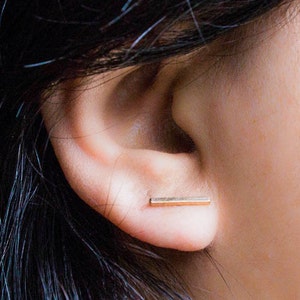 May include: A close-up of a person's ear with a small, rectangular gold stud earring. The earring is a simple bar shape, and the skin around the ear is smooth and light-toned. Dark hair frames the ear.