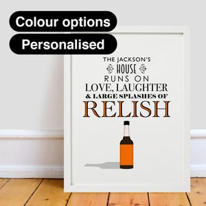 Op de afbeelding: Witte ingelijste print met zwarte tekst die "The Jackson's House Runs on Love, Laughter & Large Splashes of Relish" luidt. Onder de tekst is een bruine fles relish afgebeeld.