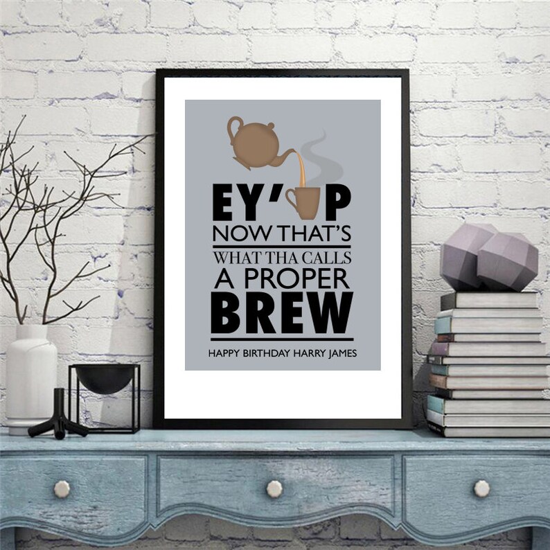 May include: A framed print with a gray background and black text that reads "Ey' up now that's what tha calls a proper brew". The text is accompanied by a cartoon image of a teapot pouring tea into a cup. The bottom of the print reads "Happy Birthday Harry James".