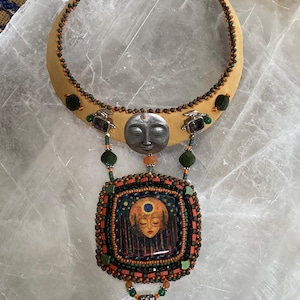 May include: A crescent-shaped collar necklace featuring a silver-toned face charm, a square pendant with a painted face surrounded by forest imagery, and a dangling iridescent gemstone. The piece is embellished with green, orange, and black beads.
