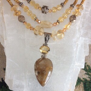 May include: A gold and silver necklace with a teardrop-shaped pendant featuring a clear quartz crystal with gold inclusions. The necklace is made with multiple strands of small, round, yellow beads.