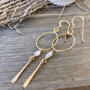 May include: Gold hoop earrings with a small pearl dangle and a long, thin gold bar hanging from the pearl.