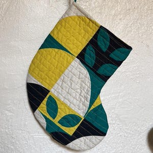 May include: A quilted Christmas stocking with a geometric pattern. The design features yellow, white, teal, and black shapes, including leaf-like motifs. The stocking is hanging on a wooden rack with gold-colored hooks.