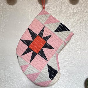 May include: A quilted Christmas stocking with a starburst design. The stocking features a pink background with white and black geometric shapes. The starburst is black with a red center. A red loop is attached for hanging.