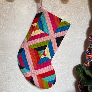 May include: A colorful Christmas stocking with a patchwork design. The stocking features a variety of colors including pink, red, blue, green, and yellow. It is hanging from a wooden rack with gold hooks. The stocking is approximately 12 inches long.