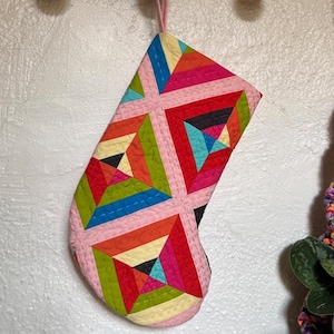 May include: A quilted Christmas stocking with a geometric pattern in pink, red, orange, yellow, green, blue, and black. The stocking is hanging from a wooden hook on a white wall. The stocking has a pink border.