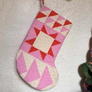 May include: Pink and white quilted Christmas stocking with a star and triangle pattern. The stocking is hanging from a wooden bar with gold hooks. The design features red, pink, and cream colors.
