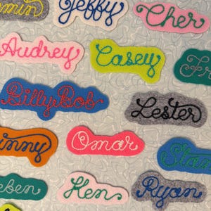 May include: A collection of felt name patches featuring embroidered cursive text in various colors including Jeffy, Cher, Audrey, Casey, BillyBob, Lester, Ginny, Omar, Stan, Ruben, Ken, Ryan, Markus, and Avon on assorted felt shapes.