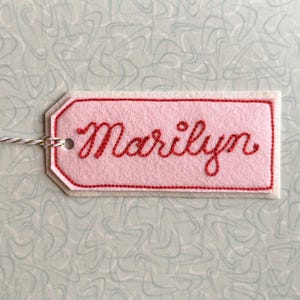May include: A pink felt tag with the name "Marilyn" embroidered in red cursive. The tag has a red border and is attached to a white and silver string. The background is a light blue pattern.