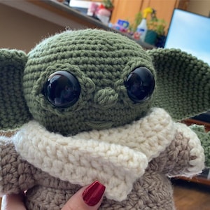 May include: A green crocheted plush toy with large black eyes and a white crocheted scarf. The toy is wearing a brown crocheted outfit. The toy is sitting in front of a television.