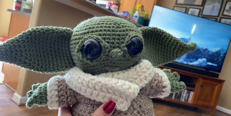May include: A green crocheted plush toy with large black eyes and a white crocheted scarf. The toy is wearing a brown crocheted outfit. The toy is sitting in front of a television.