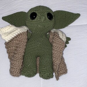 May include: A green crocheted plush toy with large eyes and a brown and cream crocheted cape. The toy is a popular character from the Star Wars franchise.
