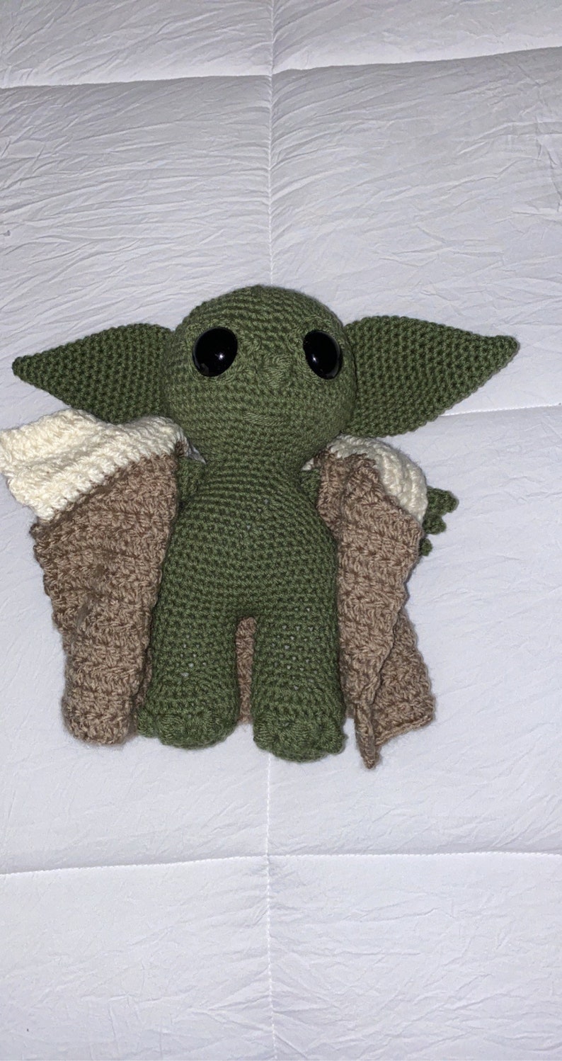 May include: A green crocheted plush toy with large eyes and a brown and cream crocheted cape. The toy is a popular character from the Star Wars franchise.