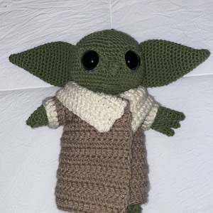 May include: A green crocheted plush toy with large black eyes, wearing a brown crocheted coat with a white crocheted collar.