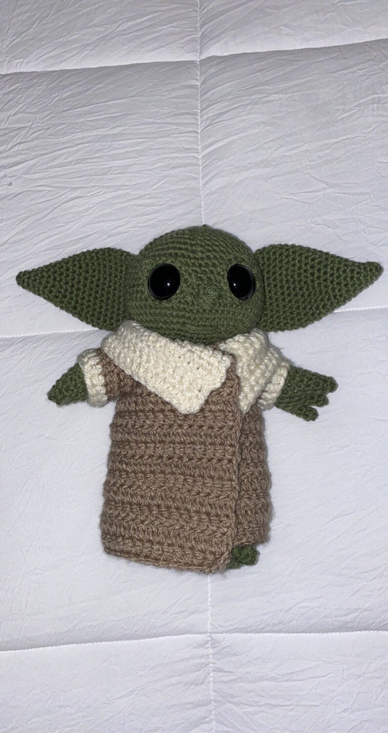 May include: A green crocheted plush toy with large black eyes, wearing a brown crocheted coat with a white crocheted collar.