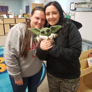 May include: Two women are smiling and holding a green crocheted stuffed toy that resembles a character from the Star Wars franchise. The toy is wearing a brown hooded robe. The women are standing in a classroom with bookshelves and a whiteboard in the background.