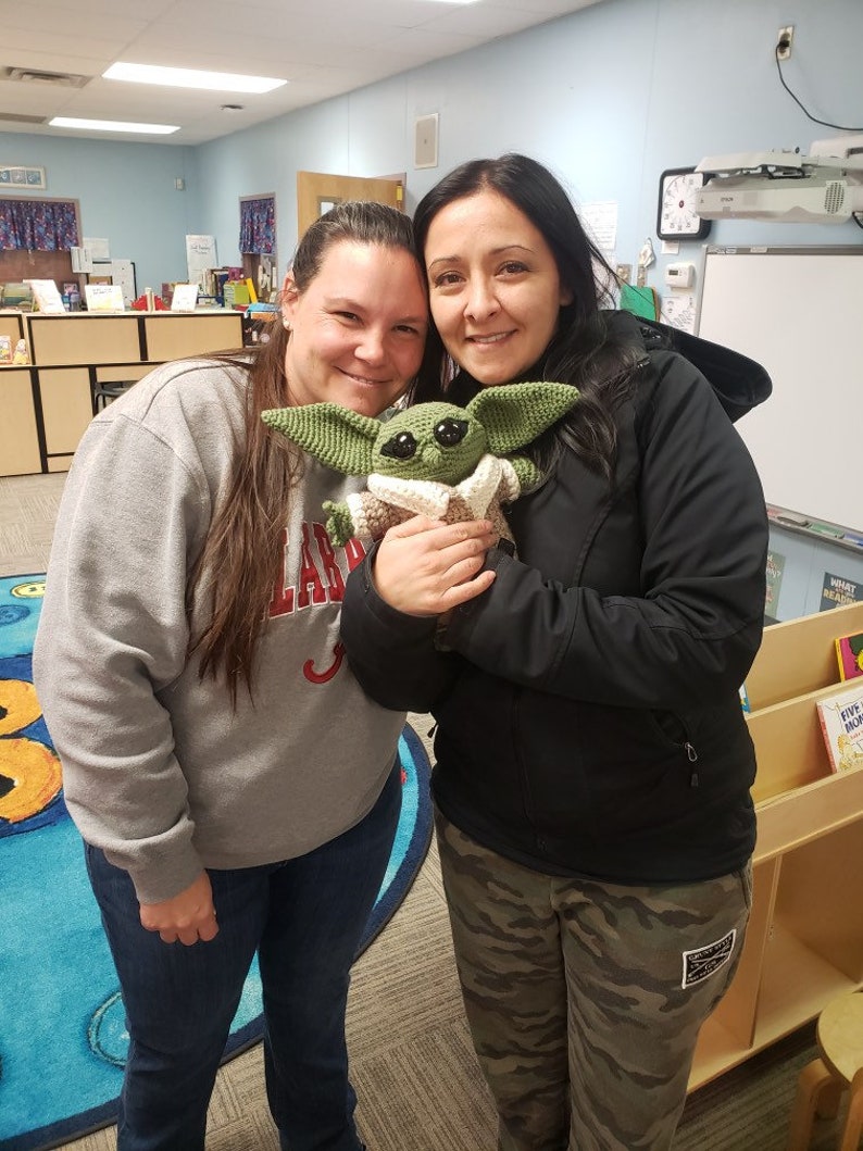 May include: Two women are smiling and holding a green crocheted stuffed toy that resembles a character from the Star Wars franchise. The toy is wearing a brown hooded robe. The women are standing in a classroom with bookshelves and a whiteboard in the background.