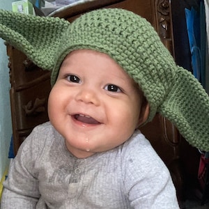 May include: A baby wearing a green crocheted hat with large, pointed ears, resembling a character from a popular science fiction franchise. The baby smiles, wearing a grey long-sleeved top.