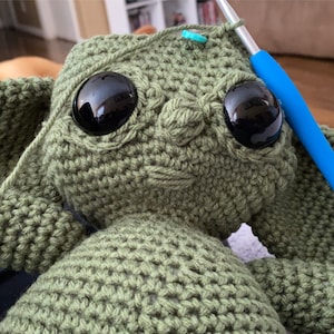 May include: A green crocheted stuffed toy with large black eyes. The toy is being worked on with a blue crochet hook.