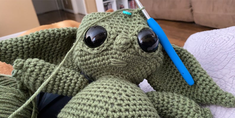 May include: A green crocheted stuffed toy with large black eyes. The toy is being worked on with a blue crochet hook.