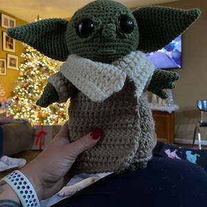 May include: A crocheted green and brown plush toy with large eyes and a white crocheted collar. The toy is being held by a person's hand.