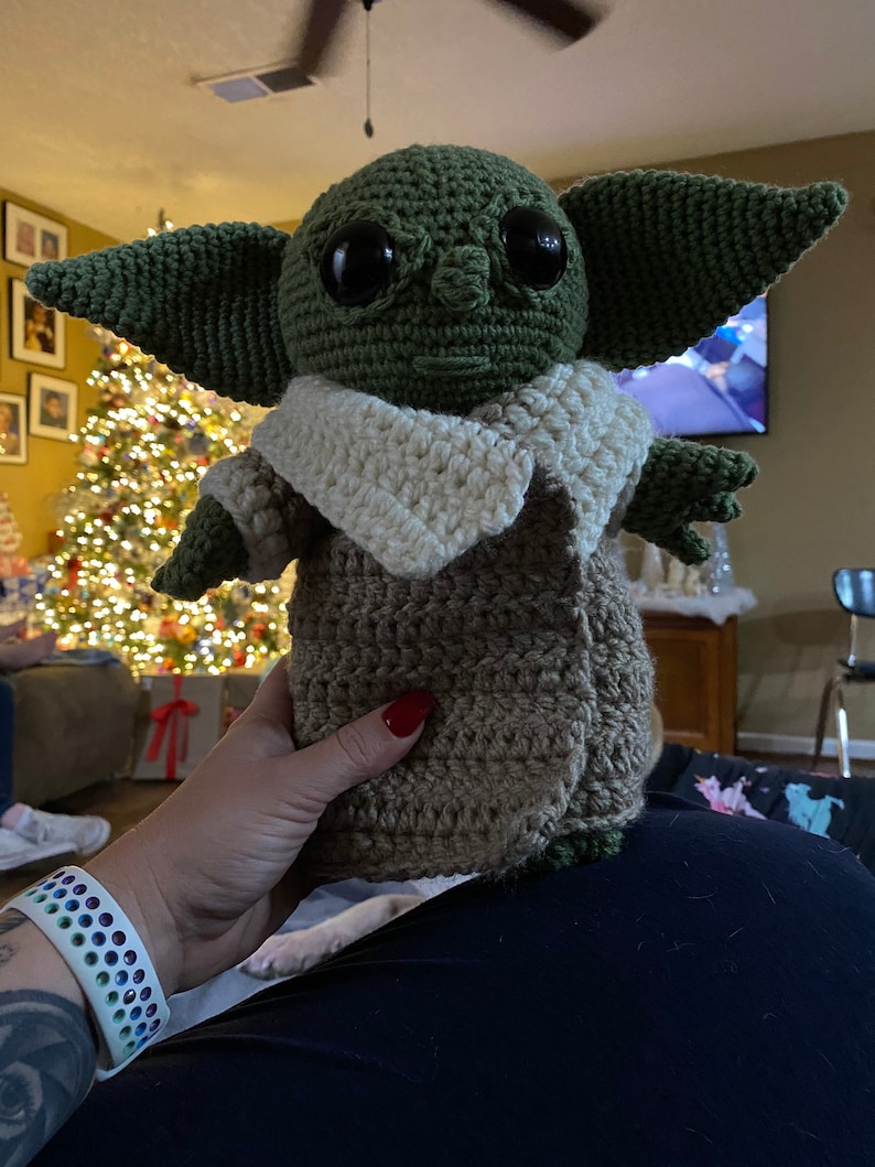 May include: A crocheted green and brown plush toy with large eyes and a white crocheted collar. The toy is being held by a person's hand.