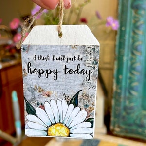 May include: A wooden tag with a jute string for hanging. The tag features the text "I think I will just be happy today" above a large white daisy with a yellow center. The background has a distressed, floral design.
