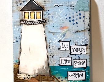 May include: A mixed media art piece on a rectangular board featuring a white lighthouse with a black roof and yellow windows. The background includes collage elements of text and blue paint. The text "Let YOUR light shine bRight" is displayed.
