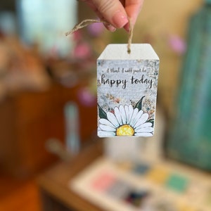 Puede incluir: Una etiqueta rectangular de madera con una ilustraci&oacute;n de margarita y el texto "I think I will just be happy today." La etiqueta tiene una parte superior blanca y una cuerda de yute para colgar. La margarita tiene p&eacute;talos blancos y un centro amarillo.
