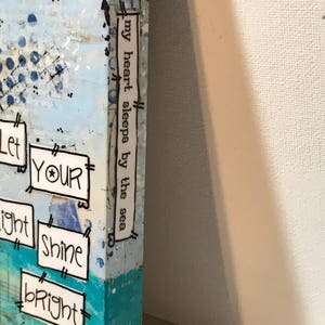 May include: A blue and white canvas painting with the text "Let your light shine bright" and "my heart sleeps by the sea" on the side.